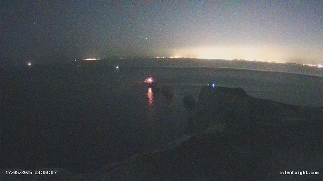 A nighttime scene on a beach showing the ocean, with reflections of distant lights on the water. The sky is clear with visible stars and a faint glow on the horizon. The timestamp indicates the date and time as May 17, 2025, at 11:00 PM.
