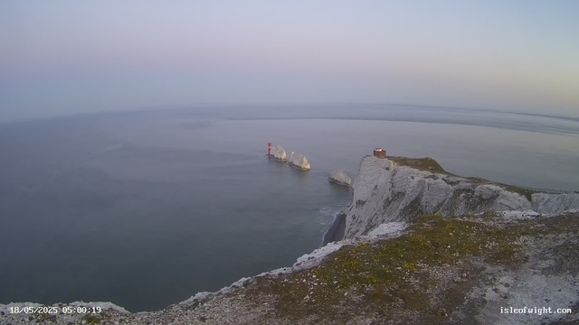 A view of chalk cliffs along a coastline with a small building on top. In the water below, there are large sea stacks, one with a red lighthouse on top. The sky is clear with a slight gradient from light blue to pale pink, indicating early morning. The image includes a timestamp in the bottom left corner, reading "18/05/2025 05:00:19," and a website URL "isleofwight.com" in the bottom right corner.