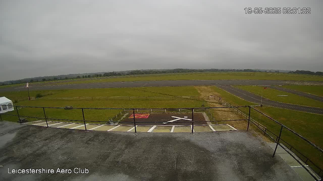 An outdoor view of a runway with a grassy area in the background. In the foreground, there is a rooftop with a railing, a helipad marked with a white "H" and a red section, and parking spaces. The sky is overcast with gray clouds. The timestamp in the top right corner reads "18-05-2025 05:01:02" and the bottom left corner has the text "Leicestershire Aero Club."