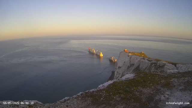 A view from a webcam showing white chalk cliffs overlooking the ocean at sunrise or sunset. A small building and a lighthouse are situated on top of the cliffs. There are a few large rock formations emerging from the water just offshore, and the sea appears calm. The image has a timestamp of May 18, 2025, at 6:00 AM.