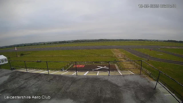 An empty parking area with designated spots, including a large white arrow painted on the ground pointing to the right. There is a grassy field extending into the distance with a runway or taxiway visible, and a cloudy sky overhead. A small sign with red and white markings can be seen in the background, indicating it is part of the Leicester Shire Aero Club premises. The timestamp in the upper right corner reads "18-05-2025 06:00:50."