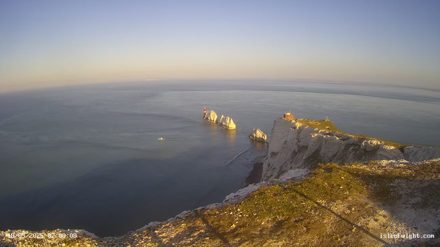 Aerial view of white chalk cliffs and rock formations extending into the sea during sunset, with a small building on the cliff. The ocean appears calm with a boat leaving a trail in the water. The sky is clear with soft sunlight illuminating the landscape.