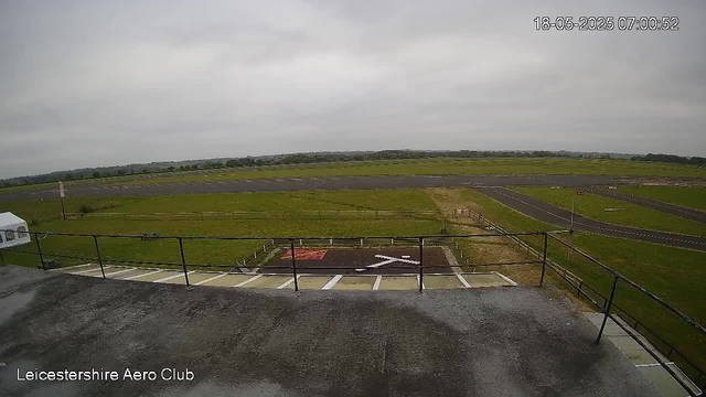 An overcast daytime view from a webcam showing an empty aircraft parking area with painted markings, a grassy airfield with taxiways, a low fence, and a slightly cloudy sky. The timestamp in the upper right corner indicates it is 7:00 AM on May 18, 2025. The bottom left corner displays the label "Leicestershire Aero Club".