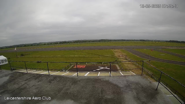 A view of an open grassy area with a small tarmac section and a taxiway, surrounded by a fence. Overcast sky with gray clouds. The scene appears to be a small airfield or airport. The image is timestamped "18-05-2025 07:59:59" and labeled "Leicestershire Aero Club" at the bottom left corner.
