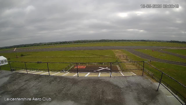 The image shows a view from a webcam overlooking an airfield with a parking area in the foreground. The parking lot has white parking lines and a helipad with a white "H" marked on it, surrounded by a small fenced area. Beyond the parking, there are grassy fields and a racetrack with a curved layout. The sky is overcast with thick gray clouds, and there are no visible people or aircraft in the scene. The timestamp in the top right corner indicates it was captured on May 18, 2025, at 9:00:49 AM. The bottom left corner displays the text "Leicestershire Aero Club."