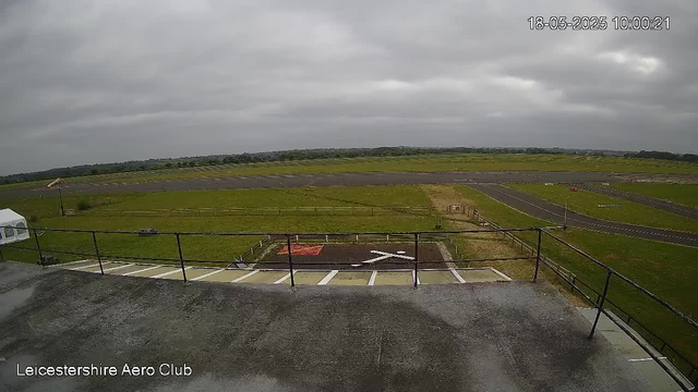 A view of an airfield with a runway and surrounding grassy area under a cloudy sky. The image appears to be taken from a webcam at the Leicester/shire Aero Club, with a timestamp showing 10:00 on May 18, 2025. There is a small parking area with marked spaces in the foreground, and a fence borders the edge of the platform from where the photo is taken.
