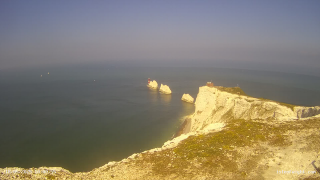 A view from a webcam shows white chalk cliffs on the right, extending into the sea. In the distance, there is a lighthouse on a small rock formation jutting out into the water, which is calm and greenish in color. The sky above is clear with a gradient from blue at the top to lighter hues near the horizon. Several small sailboats are scattered across the water. The timestamp at the bottom left indicates the date and time as 18/05/2023 at 10:00:55.