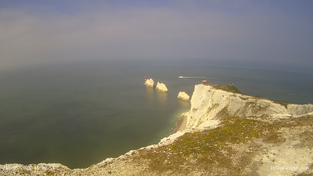 A panoramic view of white chalk cliffs extending into the ocean, with a lighthouse on a distant headland and several rock formations in the water. The sky is partly cloudy, and a small boat is visible in the distance.