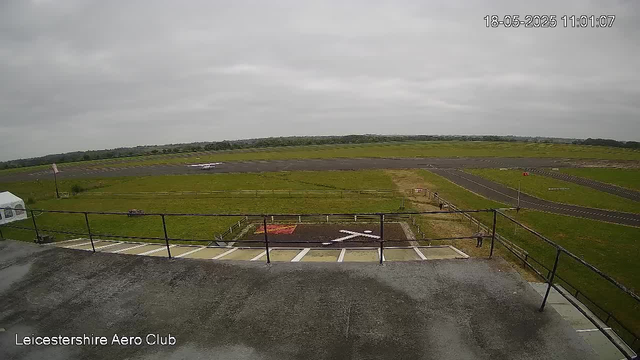The image is a view from a webcam at Leicestershire Aero Club, showing a cloudy sky over a small airfield. In the foreground, there is a rooftop with safety railings and a small helipad marked with a white "H" and a red border. Beyond the rooftop, there is a grassy area with a person standing near the edge, and a small aircraft parked on the tarmac. Runways and taxiways extend into the distance, with grassy fields surrounding the airfield. The timestamp on the top right corner indicates the date as May 18, 2025, and the time as 11:01:07.