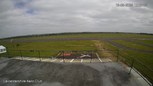 A view from a webcam at Leicesterhire Aero Club showing a runway and grassy fields under a cloudy sky. The timestamp in the top right corner is "18-05-2025 12:00:02." The foreground includes a balcony with a metal railing and some parking spots.