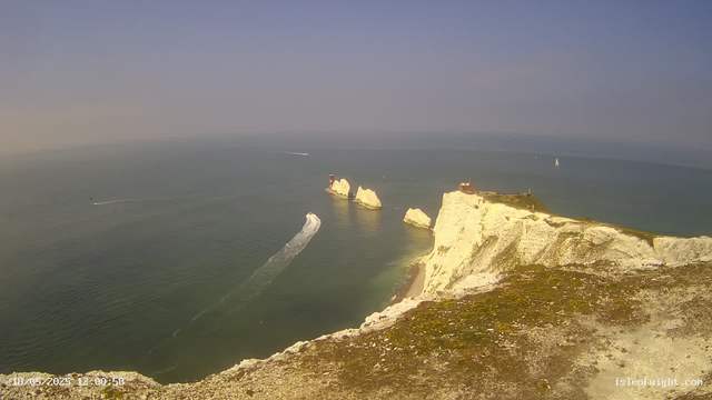A panoramic view of white chalk cliffs along the coastline, with green grass on top. A small building and a lighthouse are visible on the highest point of the cliffs. The sea below shows several boats, including a sailboat and a vessel creating a trail of wake across the water. The sky is clear with a slight haze on the horizon. The timestamp on the bottom left indicates the date and time as May 18, 2023, at 12:00:58.