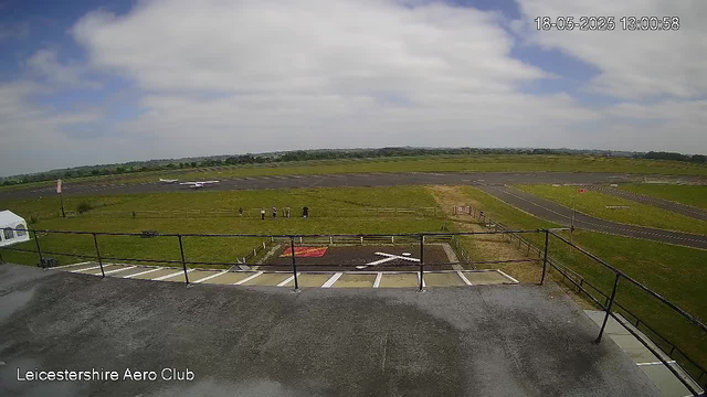An image from a webcam showing an outdoor area at Leicestershire Aero Club. In the foreground, there is a small parking area with a white vehicle partially visible on the left. Beyond the parking lot, a grassy field with a few small groups of people standing near a fence. Further back, there is a runway with a small aircraft parked on the side. The sky is partly cloudy with patches of blue sky visible. The timestamp indicates the image was captured on May 18, 2025, at 1:00:58 PM.