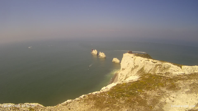 Aview of white chalk cliffs extending into the ocean with large rock formations in the water. There are small boats moving across the water, leaving white wakes behind them. The sky is clear with a faint gradient from blue to a light hue near the horizon. The date and time are displayed in the bottom left corner.