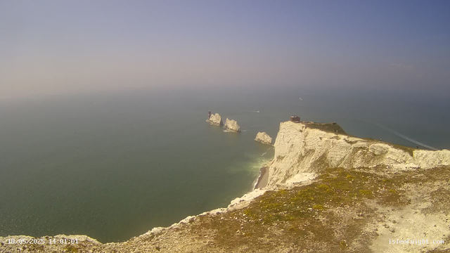 A view of white chalk cliffs along a coastline, with three prominent rock formations in the sea. The sky is clear with a gradient from light to darker blue as it meets the horizon. A small structure is visible on top of the closest cliff, and a boat is seen sailing in the distance. The camera timestamp reads "18-05-2025 14:01:01".