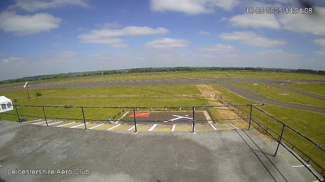 A wide view of a small airport runway under a partly cloudy sky with scattered white clouds. In the foreground, there is a parking area with a white vehicle partially visible on the left side. A black railing runs along the edge of the parking area, and on the ground beneath it, there is a helipad marked with a white "H" and a red border. The rest of the field is green, with a paved runway extending into the distance and some small white structures or objects along its sides. The timestamp on the top right corner reads "18-05-2025 14:01:08" and the bottom left corner has the label "Leicestershire Aero Club."