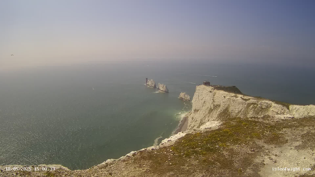 A panoramic view of a coastal landscape featuring white chalk cliffs with patches of greenery at the top. In the distance, there are several clusters of rock formations emerging from the sea. A small building or structure is situated near the edge of the cliff. The ocean appears calm with gentle waves, and a few boats are visible on the water. The sky is clear with a gradient from light to darker blue. The image also shows the timestamp "18/05/2025 15:00:33" at the bottom left corner.