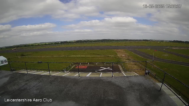 A view from a webcam showing a grassy airfield with multiple runways stretching into the distance. The sky is partly cloudy with patches of blue. In the foreground, there is a small parking area with designated parking spots and a black railing, along with a helipad marked with a white "H" and a red border. The timestamp in the upper right corner reads "18-05-2025 15:00:37." In the lower left corner, there is text that reads "Leicestershire Aero Club."