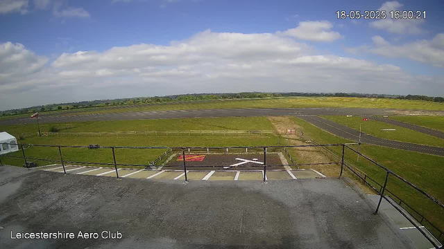 An outdoor view of Leicestershire Aero Club showing a runway and grassy areas under a partly cloudy sky. There is a helipad marked with a white "H" on a dark surface in the foreground. A small flag, possibly an aircraft tail, is visible on the left side. The image is timestamped "18-05-2025 16:00:21" in the top right corner.