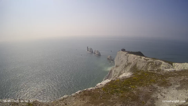 A view of the white chalk cliffs along the coastline, with the sea extending to the horizon. Several large rock formations are situated offshore, and a lighthouse is visible atop one of the distant rocks. The sky is clear with a subtle gradient from light to darker blue. The foreground shows the edge of the cliff, with some patches of moss and dirt. The timestamp and website URL are visible at the bottom left and right corners of the image.