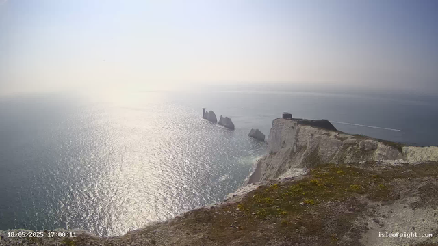 A view of white chalk cliffs overlooking the ocean, with the sunlight reflecting on the water. In the distance, there are notable rock formations emerging from the sea, and a lighthouse or similar structure sits atop the cliff. The date and time are displayed at the bottom left corner of the image.