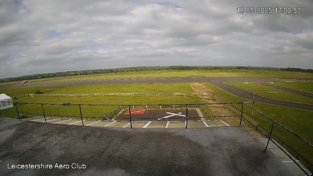 A view of an airfield with a runway stretching into the distance, surrounded by green grass and some small patches of dirt. There is a parking area with designated spaces and a small red and white wind sock on the left side. The sky is partly cloudy with patches of blue. The image is timestamped May 18, 2025, at 17:00:53, and the bottom left corner has the text "Leicestershire Aero Club."
