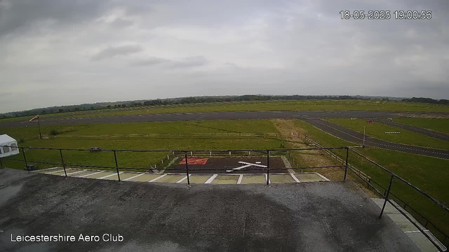 A view of an airfield with a runway extending into the distance under a cloudy sky. In the foreground, there is a balcony with a metal railing and a small helipad marked with a white H. The image is time-stamped "18-05-2025 19:00:56" in the upper right corner, and the bottom left corner has the text "Leicestershire Aero Club."