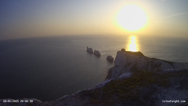 A scenic view of white chalk cliffs at sunset, with the sun low on the horizon casting a golden reflection on the calm ocean. Three prominent sea stacks rise from the water near the cliffs, and a small building or structure is visible on top of the cliffs to the right. There is a pathway or railing along the edge of the cliffs. The timestamp shows "18/05/2025 20:00:48" and the website "isleofwight.com" is displayed in the bottom right corner.