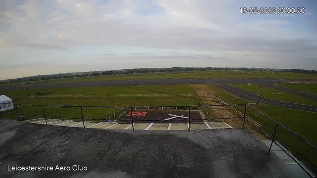 An outdoor view from a webcam shows a large open grassy area with a small paved runway or taxiway in the distance, surrounded by a fence. There is a helipad marked with an "X" and a red square. The sky is partly cloudy with patches of blue, and the timestamp reads "18-05-2025 20:00:50." The bottom left corner indicates the location as "Leicestershire Aero Club."