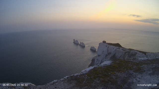 A view from a coastal cliff at sunset shows white chalk cliffs extending into the sea with large sea stacks just offshore. In the distance, there is a structure that appears to be a lighthouse or tower, and a small building is visible on top of the cliffs. The sky is partly cloudy with soft pastel colors from the setting sun, and the water below is calm. The timestamp in the bottom left corner indicates the date and time as May 18, 2025, at 21:00:02, and the bottom right has a website URL "isleofwight.com."