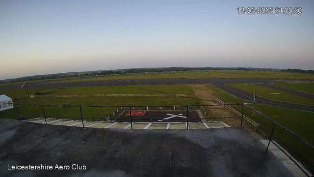 An early evening view from a webcam at Leicesterhire Aero Club showing a grassy airfield with a small runway and taxiways stretching into the distance. There is a runway with windsock on the right side, and a helipad marked with a white helicopter symbol and red border in the foreground. The sky is clear with a faint gradient from blue to light near the horizon. The date and time stamp in the upper right corner reads 18-05-2025 21:00:20.