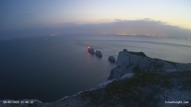 A view of a coastal cliff at dusk with a rocky shoreline extending into calm water. There are three distinctive rocks near the shore, with a small tower or lighthouse emitting a red light on one of them. A faint blue light is visible on the edge of the cliff. The sky is partly cloudy with a gradient from light to darker hues near the horizon, and distant city lights can be seen across the water. The timestamp shows "18/05/2025 22:00:32" and a website link "isleofwight.com".