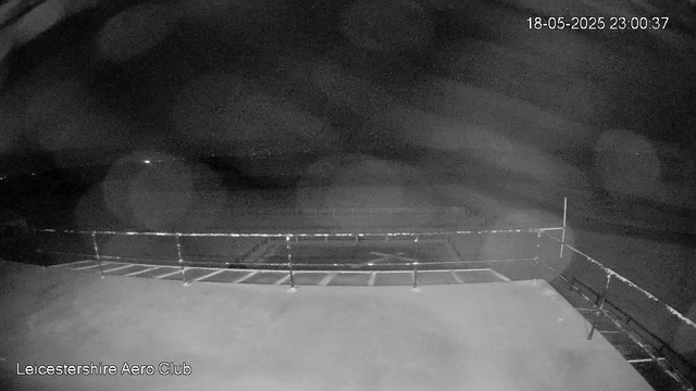 A black and white night-time image of an outdoor area with a fenced perimeter. The scene appears to be an airport or airfield, and the image is from a CCTV camera labeled "Leicestershire Aero Club" in the bottom left corner. The timestamp in the top right corner shows the date as May 18, 2025, at 11:00 PM and 37 seconds. The sky is cloudy or hazy, with no visible lights or activity.