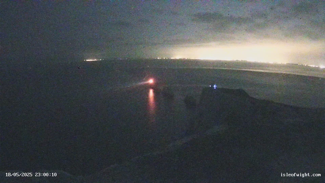 A dark sky over the ocean at night, with a faint glow of city lights visible on the horizon. There are a few small lights on the water, possibly from boats or buoys. A bright red light near the water's surface reflects on the ocean, and a distant light is visible along the horizon. The scene appears to be captured from the window of an aircraft or a high vantage point.
