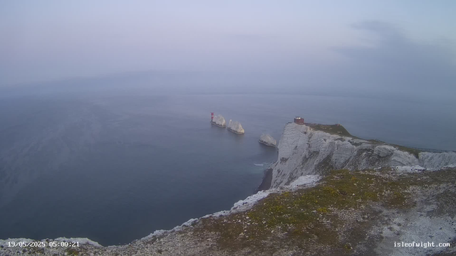 A view of white chalk cliffs overlooking the ocean at dawn, with rugged rocks protruding from the water and a lighthouse perched on a distant rock formation. The sky is overcast with a soft, muted light.