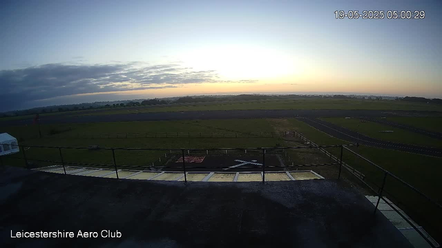 A view from a webcam showing an open field with a runway and a helipad, under a partly cloudy sky during sunrise or sunset. The timestamp in the top right corner reads 19-05-2025 05:00:29, and the bottom left corner indicates "Leicestershire Aero Club."