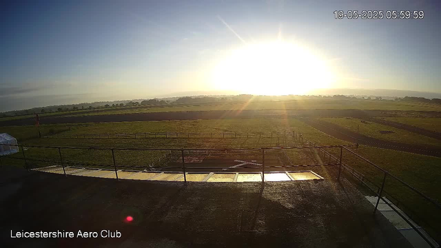 A wide outdoor view of a grassy field with several fenced areas, taken at sunrise or sunset. The sky is mostly clear with the sun low on the horizon, casting bright light and long shadows across the scene. There are some distant trees or low hills in the background. In the bottom left corner, the text "Leicestershire Aero Club" appears. The top right corner displays the date and time: 19-05-2025 05:59:59.