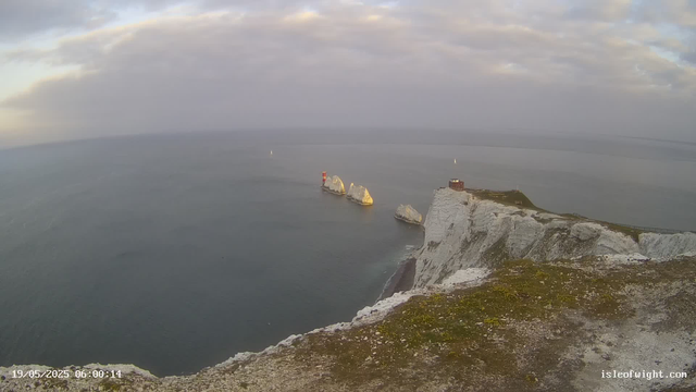 An aerial view of white chalk cliffs overlooking the sea at dawn. Three large rock formations are visible in the water near the cliffs, with one topped by a small red and white lighthouse. A building is situated on the edge of the cliffs, connected by a narrow path. The sky is partly cloudy with soft light. The image has a timestamp of May 19, 2025, at 06:00:14, and a website URL in the bottom right corner.