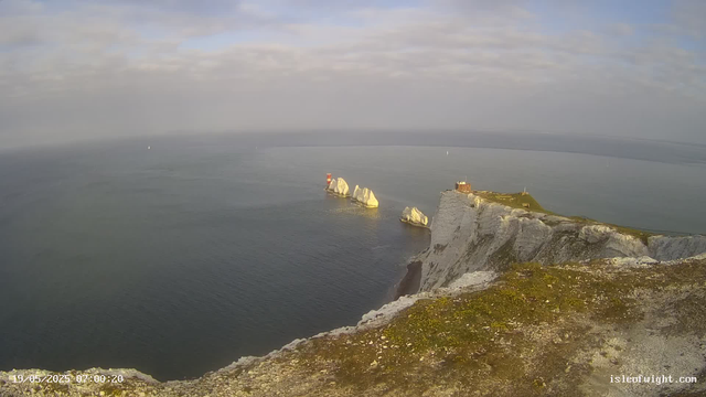 A view of white chalk cliffs extending into the sea with several large rocks and an island with a small building and a lighthouse on top. The sky is partly cloudy and calm water surrounds the cliffs.