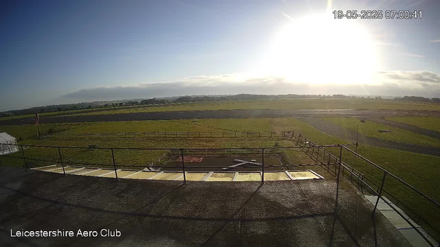 An outdoor view from a webcam shows a bright, sunny morning at Leicesterhire Aero Club. In the foreground, there is a rooftop with safety railings and some structure possibly related to aircraft maintenance or parking. Beyond, there are open grassy fields with some paved paths and runways. The horizon features a low cloud layer and distant buildings or trees. The sky is mostly clear with the sun shining brightly, creating glare in the image. The timestamp indicates it is early morning on May 19, 2025.