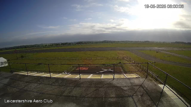 An outdoor view from a webcam showing a runway and grassy fields under a partly cloudy sky. The sun is shining brightly near the horizon, creating a glare. There is a fenced area with a helipad marked with an "H" in the foreground. The image is timestamped "19-05-2025 08:00:06" and has the label "Leicestershire Aero Club" at the bottom left corner.