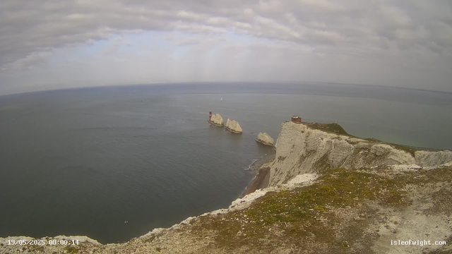 A view from a webcam showing white chalk cliffs overlooking the sea. There are three prominent white rock formations in the water near the cliffs, with a red and white lighthouse on top of one of the rocks. The sky above is mostly cloudy with some patches of blue, and the water appears calm. A small boat is visible in the distance on the water, and the image has a timestamp indicating it was taken on May 19, 2025, at 08:00:14.