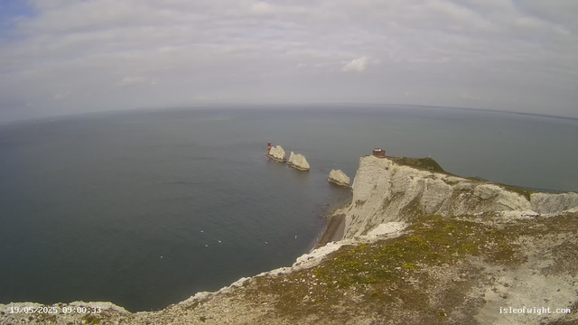 A panoramic view of white chalk cliffs overlooking a calm sea under a cloudy sky. Several large rock formations and a small lighthouse are visible in the water near the base of the cliffs, with a structure on top of the cliff to the right. The scene appears to be taken from a high vantage point on the cliffs.