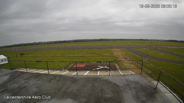A view from a webcam at LeicesterShire Aero Club showing a cloudy sky over a runways and grassy area. There are cars parked in the foreground, a small fence, and a tarmac path with directional markings. The time and date are displayed in the top right corner as "19-05-2025 09:00:18."