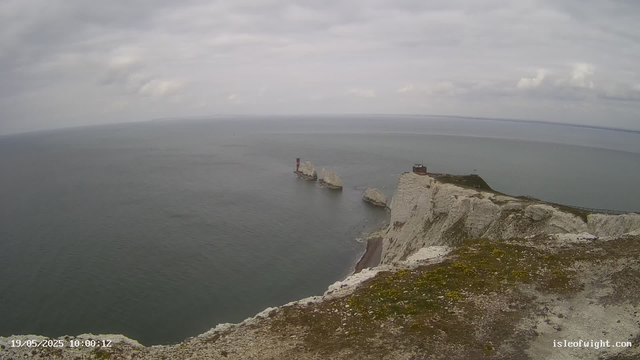 A view of white chalk cliffs approaching the ocean, with a narrow pathway along the edge. There are several large rocks in the water near the base of the cliffs, and a small building or structure on top of the cliff, overlooking the sea. The sky is cloudy with overcast conditions.