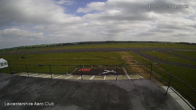 A wide view of an open grass airfield under a partly cloudy sky. In the foreground, there is a concrete platform with a black railing, and a helicopter pad with a white "H" marked on it. To the left, part of a white helicopter is visible. The background shows a stretch of green fields, runways, and a distant horizon. The timestamp indicates the date as May 19, 2025, at 11:00 am. The text "Leicestershire Aero Club" is displayed at the bottom left corner.