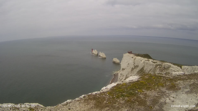 A view of white chalk cliffs extending into the ocean with several large rock formations emerging from the water. The sky is overcast with gray clouds, and a small building is visible atop the cliffs. The date and time stamp in the bottom left corner shows "19/05/2025 11:01:05."