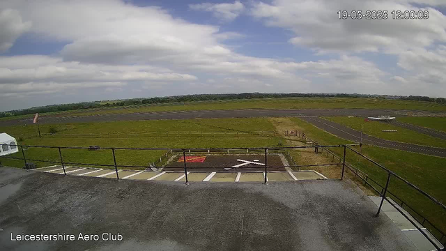 An outdoor view of an airfield under a partly cloudy sky, with a small airplane taxiing on the runway in the distance. The image includes a parking area with marked spaces and a railing in the foreground. The timestamp in the upper right corner shows the date and time as May 19, 2025, at 12:00:09. The bottom left corner has the text "Leicestershire Aero Club."