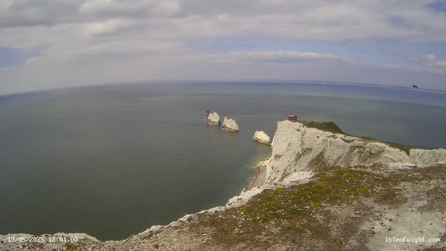 A view of the white chalk cliffs of Étretat, France, extending into the ocean. Three large rock formations, including a prominent arch and a lighthouse, stand in the water near the cliffs. The sky is partly cloudy with a mix of blue and gray clouds. A bird is flying in the distance above the water. The photo is timestamped "19/05/2023 12:01:00" and the website "isleofwight.com" is visible in the bottom right corner.