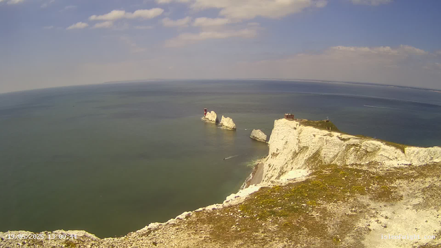 A view from a webcam showing tall, white chalk cliffs extending into the sea, with a small building and a lighthouse on top of the cliffs. In the background, there are large white rock formations in the water. The ocean appears calm and greenish, with a boat leaving a trail of white wake. The sky is partly cloudy with some scattered clouds.