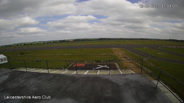 An outdoor scene showing an airfield with a large grassy area, a runway, and a few scattered structures. The sky is partly cloudy with patches of blue, and the image appears to be taken from a webcam mounted on a building. The bottom left corner has the text "Leicestershire Aero Club," and the top right corner displays the date and time: 19-05-2025 13:01:07.